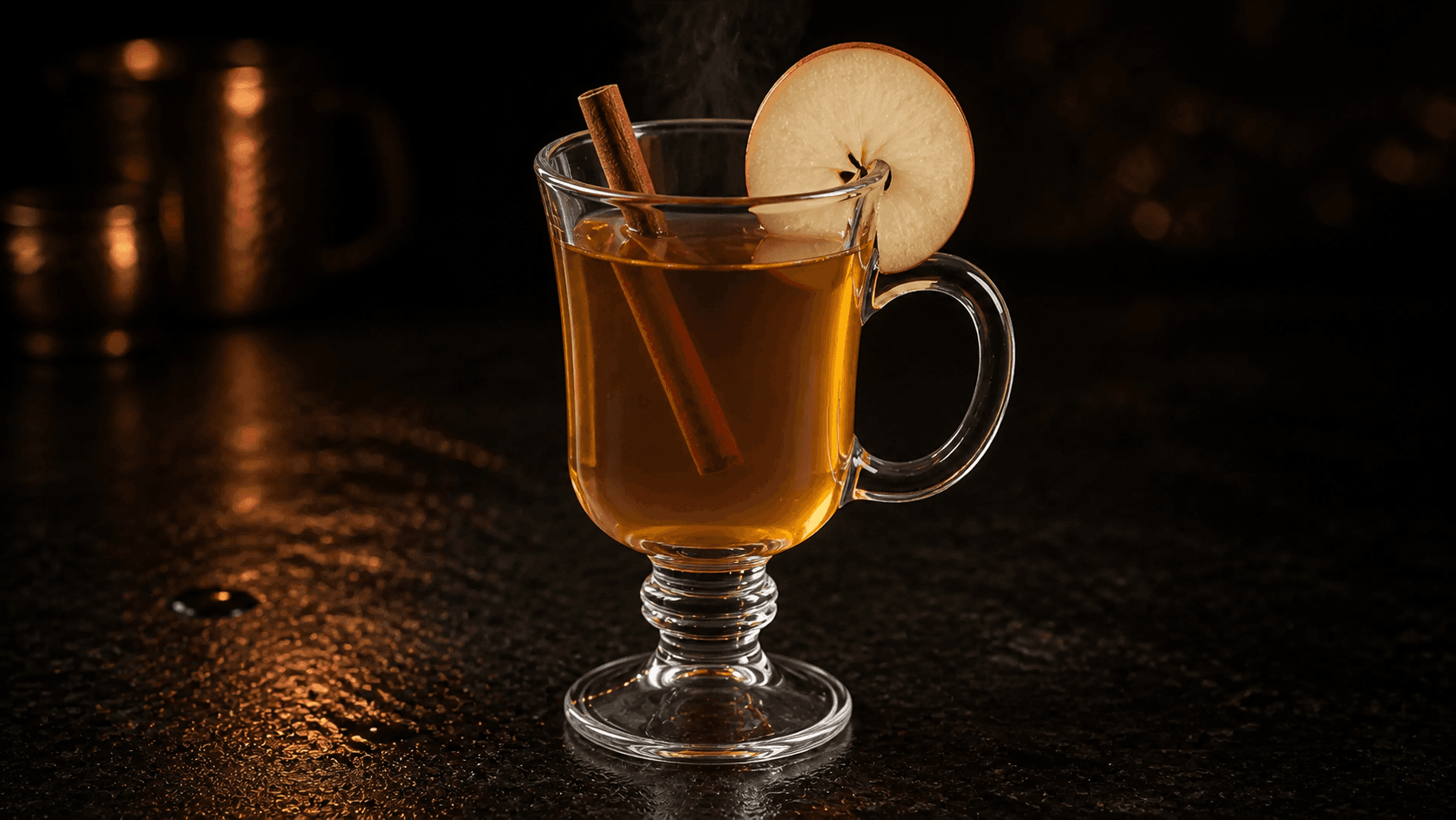 Varm äppel-toddy i ett värmetåligt glas med klar bärnstensfärgad drink, ånga, en tunn äppelskiva över kanten och en kanelstång.