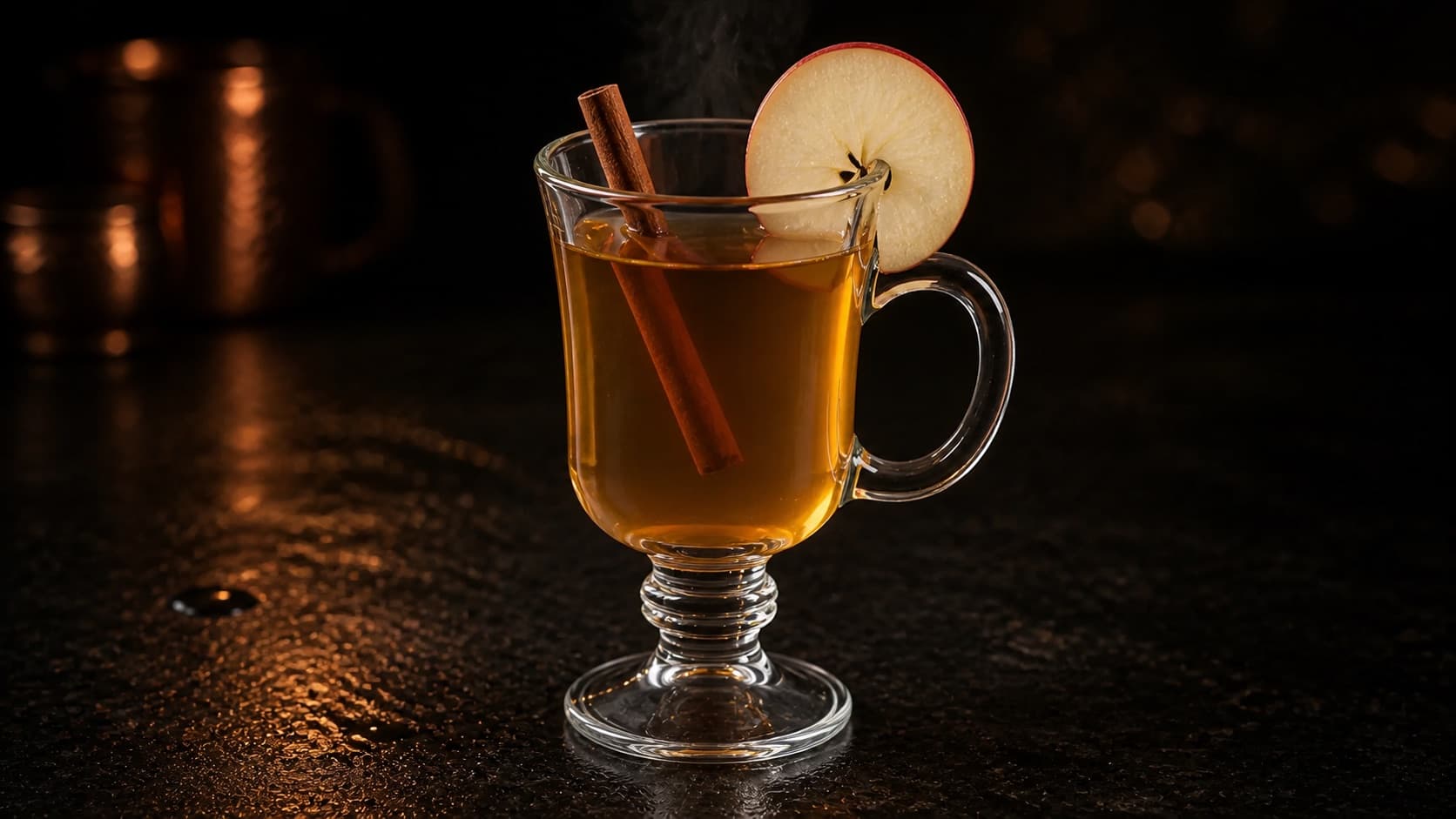 Varm äppel-toddy i ett värmetåligt glas med klar bärnstensfärgad drink, ånga, en tunn äppelskiva över kanten och en kanelstång.